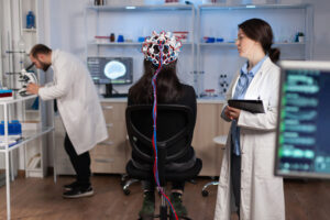 Back view of woman wearing headset for brain scan in neurology laboratory, neuro science. Nervous system analysed by team of scientists. Medical scientist looking through microscope in background developing treatment.