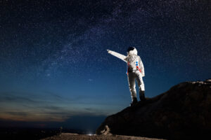 Full length of cosmonaut standing on top of rocky hill and pointing at stars. Space traveler wearing white space suit while looking at fantastic night sky. Concept of galaxy, human in space, milky way